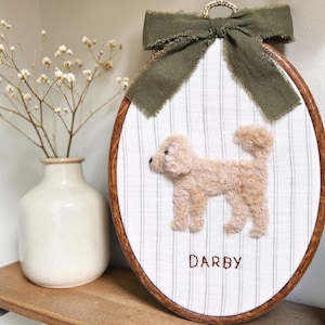 May include: Oval-shaped, framed embroidery featuring a tan dog on a striped background. The dog's name, "DARBY," is stitched below. A green bow and a gold ring are at the top. The frame is made of wood. A vase with white flowers is in the background.