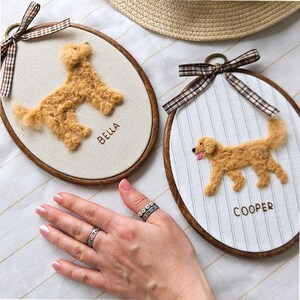 May include: Two oval embroidery hoops with brown wooden frames. One features a tan dog with the name "Bella", the other a golden retriever with the name "Cooper". Both have brown and white plaid ribbon and are on a white striped background.
