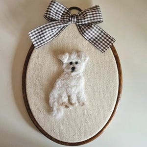Può includere: Un telaio da ricamo ovale in legno contenente uno sfondo in tessuto color crema con una figura di cane bianco realizzata con infeltrimento ad ago. Un fiocco in nastro a quadretti bianco e nero è fissato sulla parte superiore del telaio.