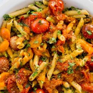 Puede incluir: Primer plano de un plato de pasta con penne, tomates cherry y hierbas. La pasta está cubierta con una salsa y adornada con hierbas frescas. El plato se sirve en un cuenco blanco, mostrando los colores vibrantes de los ingredientes.
