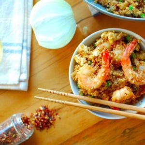 May include: A bowl of shrimp fried quinoa with chopsticks, a jar of red pepper flakes, and a white onion on a wooden table. The dish is garnished with green peas and shrimp.