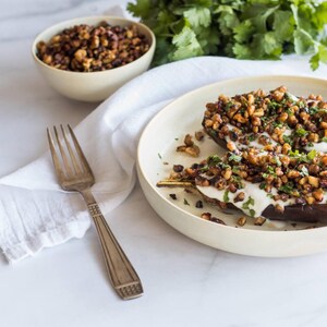 Peut inclure: Moitiés d'aubergines rôties garnies d'une sauce blanche crémeuse, de noix brunes grillées et d'herbes vertes hachées sur une assiette ronde blanc cassé, accompagnées d'une fourchette en métal et d'un petit bol de noix grillées.