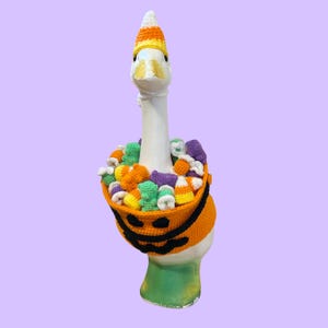 May include: A white goose figurine wearing a crocheted candy corn hat and a crocheted orange pumpkin bucket around its neck. The bucket is filled with various small, colorful crocheted candies, including shapes resembling gummy bears and candy corn.