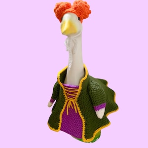 May include: A white goose figurine wearing a crocheted outfit consisting of a dark green coat with yellow trim, a purple bodice with yellow lace-up detailing, and a bright orange textured wig on its head, featuring a white crocheted neck accent.