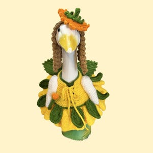 May include: A white goose figurine wearing a crocheted yellow dress with green leaf accents and a matching orange flower headpiece. The goose has brown yarn hair styled in braids and is holding a small orange flower in its wing.