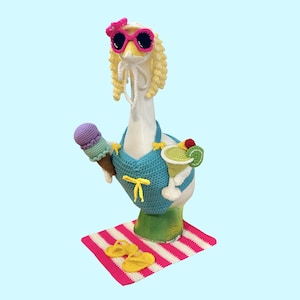 May include: A whimsical goose figurine, crocheted with a summer theme. The goose wears a blue bikini, pink sunglasses, and a yellow bow. It holds an ice cream cone and a cocktail. A pink and white striped mat and yellow flip-flops complete the beach scene.