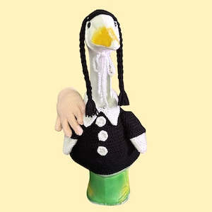 Könnte beinhalten: Eine weiße Gänsefigur, die ein schwarzes gehäkeltes Kleid mit weißem Kragen und drei weißen Knöpfen trägt. Die Gans hat zwei lange, schwarze, geflochtene Garnsträhnen am Kopf und ein kleines, blasses, gehäkeltes Handaccessoire an der Seite befestigt.