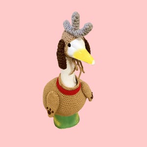 May include: A white goose figurine wearing a tan crocheted jumper with a red collar and matching tan crocheted hat. The hat features long brown ear-like flaps, a grey three-pronged crest on top, and dangling tan strings under the yellow beak.