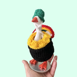 May include: A white ceramic goose figurine wearing a green crocheted hat, a yellow crocheted collar, and a black crocheted body cover. A rainbow-colored crocheted piece is attached to the back of the goose, which has an orange beak and feet.