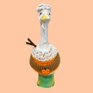 以下が含まれることがあります： 白い陶器のガチョウの置物。紐付きの白いかぎ針編みの帽子、白とオレンジの質感のある襟、小さなオレンジ色のカボチャのアップリケと2本の茶色の棒状の突起が付いた茶色のリブ編みのセーターを着用しています。