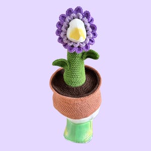 May include: A whimsical, handmade crochet flower in a terracotta pot. The flower has a purple petal head, a white and yellow beak, green stem and leaves. The pot is brown and orange, set against a light purple background.