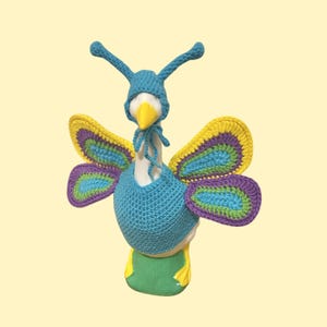 May include: A white goose figurine wearing a blue crocheted outfit consisting of a body cover and a hat with two antennae. Attached to the back are four large crocheted butterfly wings featuring concentric layers of yellow, purple, blue, and green.