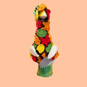 Puede incluir: Una figura de ganso de cerámica que lleva un suéter de ganchillo marrón decorado con hojas de otoño coloridas en tonos rojo, naranja, amarillo y verde. El ganso sostiene un pequeño rastrillo de ganchillo gris en sus alas blancas y tiene un pico amarillo.