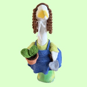 May include: A whimsical, handmade goose figurine dressed as a gardener. The goose wears a blue crochet apron, a green and yellow shirt, and a brown wig. It holds a small potted plant and a gray trowel, all crafted from yarn.