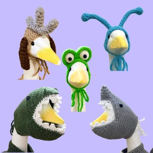 May include: Five crochet hats for geese, including a brown and tan hat with gray horns, a bright blue hat with antennae, a green frog hat with large eyes, a dark green dinosaur hat with white teeth, and a gray shark hat with white teeth.