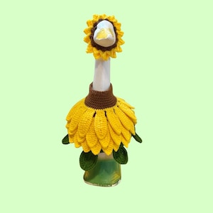 May include: A white goose figurine wearing a crocheted sunflower costume. The outfit includes a yellow sunflower headpiece around the neck and a brown and yellow sunflower petal body cover with green leaves attached at the bottom edge.