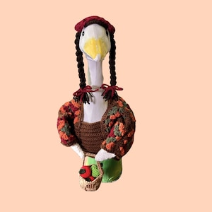 May include: A white goose figurine wearing a crocheted dark red beret, two long dark brown yarn braids, and a brown crocheted top with multicoloured granny square sleeves. The goose holds a small tan crocheted basket containing red and green fruit.