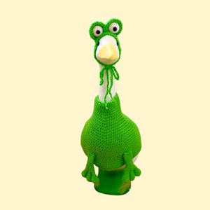 May include: A white goose wearing a green crocheted frog costume. The costume includes a frog hat with large eyes and a green body that covers the goose's body. The goose's yellow beak is visible. The background is a solid light yellow.