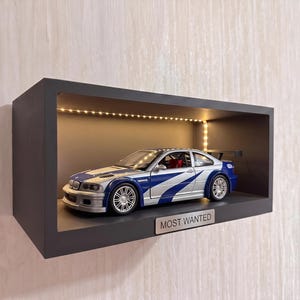 May include: A silver and blue BMW M3 GTR diecast model car displayed inside a rectangular black wall-mounted shelf with integrated LED lighting. A metal plaque with the text MOST WANTED is attached to the front edge of the shelf frame.