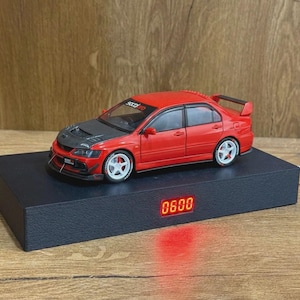 May include: A red and black scale model sedan with white wheels and a rear spoiler, positioned on a rectangular black display base featuring a digital red LED clock display showing the numbers 0600 on the front face of the base.