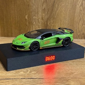 May include: A lime green and black scale model sports car featuring the number 63 on the bonnet and side, resting on a rectangular black base with a digital red LED display showing the time 06:00 and a white power cord extending from the side.