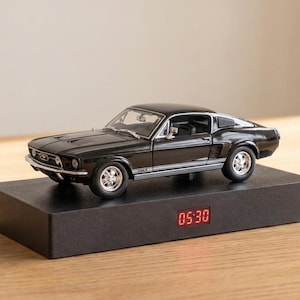 Puede incluir: Un coche a escala Ford Mustang negro se sitúa sobre una base rectangular negra que incluye un reloj digital que muestra la hora 05:30 en números LED rojos. El coche tiene ruedas plateadas y una carrocería de estilo fastback clásico.