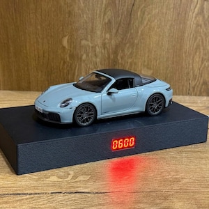 Pode incluir: Um modelo de carro Porsche 911 Targa azul claro com teto preto e rodas escuras, exibido em uma base retangular preta de levitação magnética com um display digital LED vermelho mostrando o número 0600 na parte frontal da base.