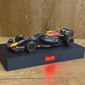 May include: A miniature Oracle Red Bull Racing Formula 1 car model in black, blue, and yellow, featuring sponsor logos, resting on a rectangular black display base with a digital red LED clock display showing the numbers 0600 on the front side.