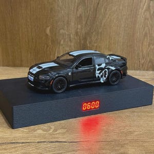 May include: A black toy car with white racing stripes and a white graphic on the side, positioned on a rectangular black base featuring a digital display showing the red numbers 0600. The base has a white power cord extending from the side.