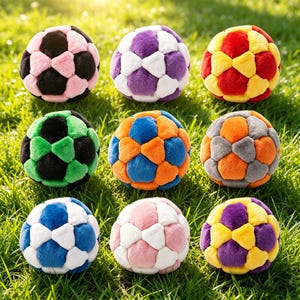Puede incluir: Nueve balones de fútbol de peluche en varios colores, incluyendo rosa, morado, verde, azul, naranja y gris. Cada balón tiene un diseño clásico de pentágonos y hexágonos. Los balones están colocados sobre hierba verde.