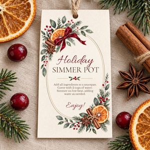 May include: A rectangular cream-coloured tag with a jute string, featuring a watercolour illustration of orange slices, cinnamon sticks, star anise, and pine branches. The text reads: Holiday SIMMER POT, instructions, and Enjoy!.