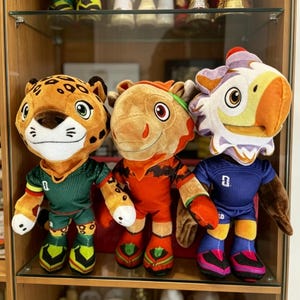 Könnte beinhalten: Drei Plüschtiere stehen nebeneinander. Links ein Leopard im grünen Trikot, in der Mitte ein beiges Wesen im orangefarbenen Trikot und rechts eine vogelartige Figur im blauen Trikot. Jedes Spielzeug weist gestickte Details im Gesicht auf.