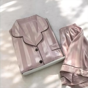 May include: A light pink striped pajama set. The top has a notched collar, a breast pocket, and dark brown buttons. The set includes a long-sleeved shirt and matching trousers. The edges of the collar, pocket, and cuffs are trimmed with dark brown.
