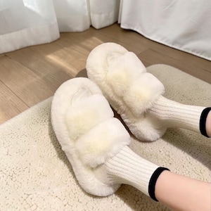 May include: A pair of off-white faux fur slides featuring two wide, plush straps across the top. The slides are worn with cream-colored, ribbed knit socks that have a thin black band at the top edge, providing a soft and textured appearance.