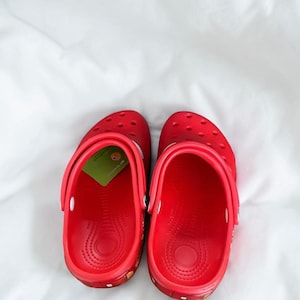 May include: A pair of bright red slip-on clogs featuring a perforated toe box, a pivoting heel strap, and a textured footbed. A small green rectangular tag with a circular graphic is tucked inside the left shoe, visible near the heel area.