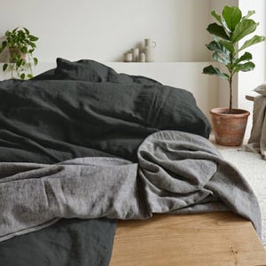 Könnte beinhalten: Ein Bett mit dunkelgrauer Leinenbettwäsche und einer hellgrauen Decke am Fußende. Das Bett steht auf einer Holzoberfläche, im Hintergrund eine Topfpflanze. Der Raum hat eine neutrale Farbpalette.