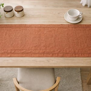 May include: A wooden dining table with a rust-colored linen table runner. A white teacup and saucer sit on the table, along with two small jars with wooden lids. A potted plant and a woven basket are also visible.