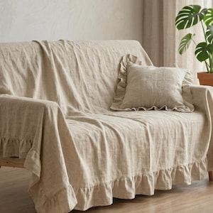 Op de afbeelding: Een beige linnen bankhoes met een gefranjerde rand en bijpassend kussen. De hoes bedekt een bank met houten frame, waardoor een zachte, getextureerde uitstraling ontstaat. Het neutrale kleurenpalet past bij het natuurlijke licht en de omringende inrichting.