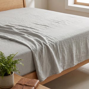 May include: A light grey bed sheet with a visible woven texture draped over a mattress. The fabric has a soft, slightly wrinkled appearance and covers the top surface of the bed, extending down the side to reveal the natural grain of the wooden frame.