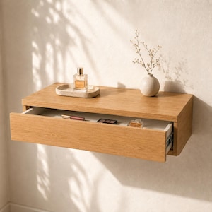 May include: A light oak floating wall shelf with a single pull-out drawer. The top surface holds a rectangular stone tray with a glass perfume bottle and a small round vase with dried stems. The open drawer contains a lipstick and palette.