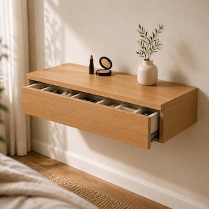 May include: A light oak floating wall shelf featuring a single wide drawer with multiple internal compartments. The top surface holds a small white ceramic vase with green foliage, a black lipstick tube, and a round black compact mirror.