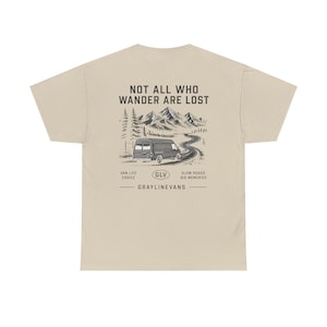 Può includere: T-shirt beige con un design grafico raffigurante un furgone che percorre una strada tortuosa tra le montagne. Il testo recita "NOT ALL WHO WANDER ARE LOST" e "VAN LIFE CHOICE, SLOW ROADS BIG MEMORIES, GRAYLINEVANS".