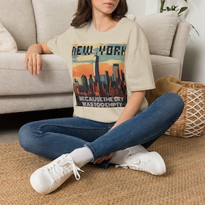 Może przedstawiać: Beżowy t-shirt z krótkim rękawem z nadrukiem graficznym przedstawiającym panoramę Nowego Jorku o zachodzie słońca. Napis NEW YORK znajduje się u góry grafiki, a napis BECAUSE THE SKY WAS TOO EMPTY znajduje się na dole.