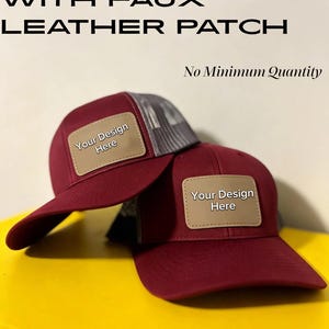May include: Two maroon trucker hats with grey mesh backs and tan faux leather patches on the front. The patches feature the text "Your Design Here". The image includes the text "CUSTOM HAT WITH FAUX LEATHER PATCH" and "No Minimum Quantity".
