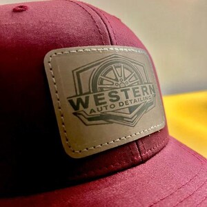 May include: A maroon baseball cap featuring a rectangular tan leather patch stitched onto the front. The patch displays a dark graphic of a car wheel inside a geometric frame with the text WESTERN AUTO DETAILING printed in a bold, dark font.