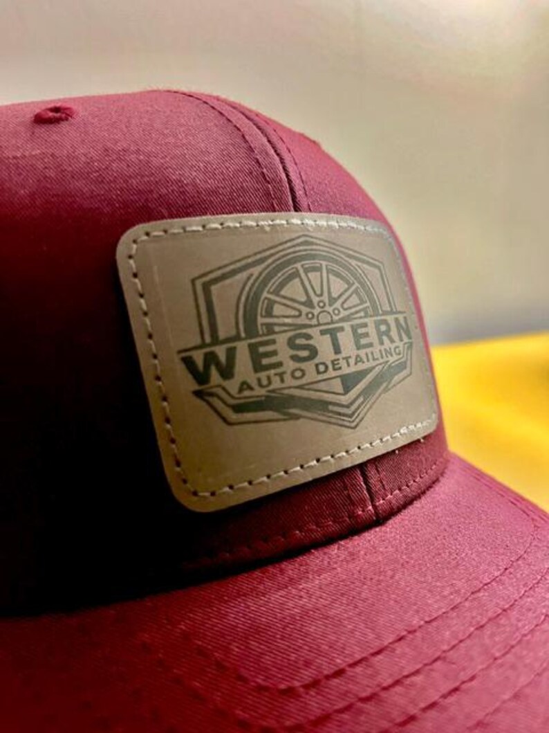 May include: A maroon baseball cap featuring a rectangular tan leather patch stitched onto the front. The patch displays a dark graphic of a car wheel inside a geometric frame with the text WESTERN AUTO DETAILING printed in a bold, dark font.