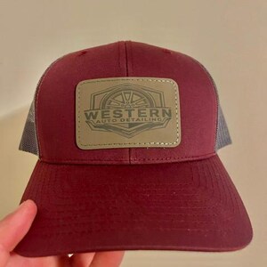 May include: A maroon baseball cap with a grey mesh back and a rectangular patch on the front. The patch is tan with a dark brown graphic of a wheel and the text WESTERN AUTO DETAILING. The cap features a curved brim and stitched detailing.