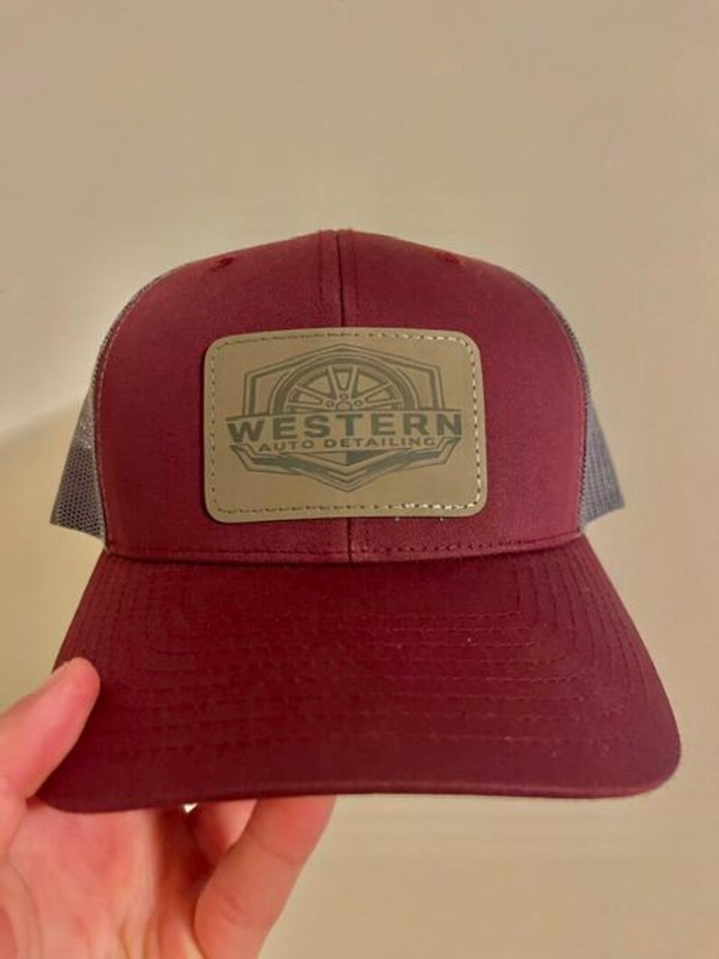 May include: A maroon baseball cap with a grey mesh back and a rectangular patch on the front. The patch is tan with a dark brown graphic of a wheel and the text WESTERN AUTO DETAILING. The cap features a curved brim and stitched detailing.