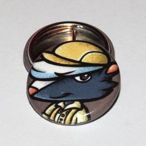 May include: A round button with a cartoon image of an animal wearing a yellow hat. The animal has a white stripe on its head, blue fur, and a brown nose. The button is on a white background.