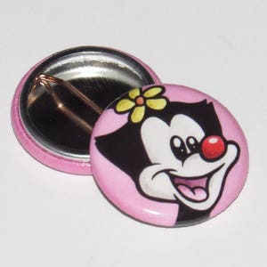 May include: Two pink pinback buttons. One button features a cartoon character with black fur, a red nose, and a yellow flower in her hair. The other button is pink with a silver metal back and a safety pin.